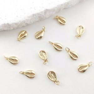 Könnte beinhalten: Zehn goldfarbene Muschel-Anhänger, verstreut auf einer weißen Oberfläche. Die Anhänger sind klein und zart, mit einem detaillierten Muschel-Design.