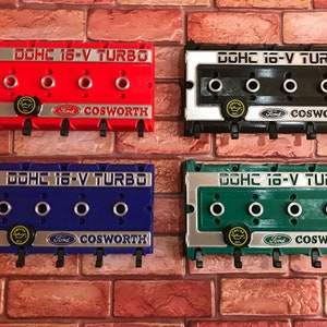 May include: Four keychains shaped like engine valve covers. Each keychain is a different colour: red, black, blue, and green. The keychains have the text "DOHC 16-V TURBO" and "COSWORTH" on them.
