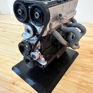 May include: A 3D printed model of a car engine with a black base and silver and gray details. The engine has a belt and pulley system and a set of exhaust manifolds.