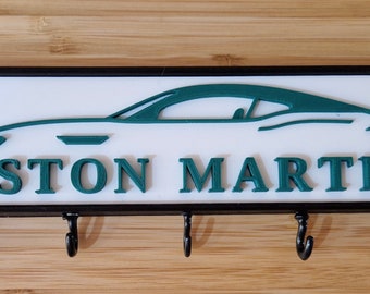 May include: A white key holder with a black frame and four hooks. The key holder features a green silhouette of an Aston Martin car and the words "ASTON MARTIN" in green letters.