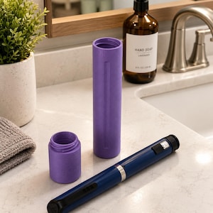 May include: A purple cylindrical protective case for an injection pen, consisting of a tall main body and a shorter screw-on cap. The case has a textured matte finish. A dark blue injection pen with a black clip and a digital display is placed nearby.