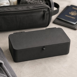 May include: A rectangular matte black storage box with a hinged lid and a small central latch. The box features a smooth, solid-colored exterior surface and a clean, simple design, providing a compact container for organizing various small items.
