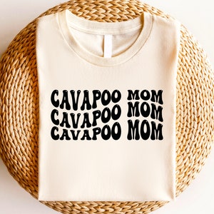 May include: A cream-colored t-shirt with the text "CAVAPOO MOM" repeated three times in a retro font.