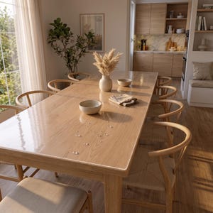 May include: A rectangular wooden dining table covered with a clear protective plastic sheet featuring scattered water droplets. The table is surrounded by light-colored wooden chairs with woven seats, and holds a ceramic vase with dried pampas grass.