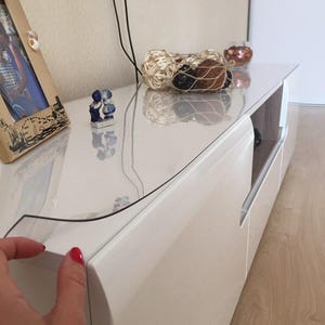 May include: A clear, flexible protective plastic cover sheet placed over the top surface of a white, glossy media console. The rectangular cover is transparent, allowing the white finish of the furniture to remain visible underneath.