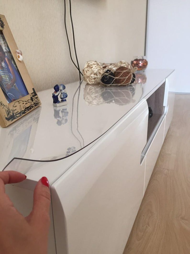 May include: A clear, flexible protective plastic cover sheet placed over the top surface of a white, glossy media console. The rectangular cover is transparent, allowing the white finish of the furniture to remain visible underneath.