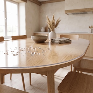 May include: A clear, oval-shaped protective table cover made of smooth plastic, featuring a glossy finish with visible water droplets scattered across the surface, resting on a light-toned wooden dining table with rounded edges and matching chairs.