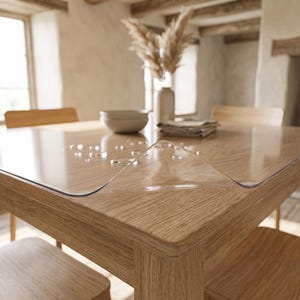 Peut inclure: Une protection de table transparente, flexible et claire, posée à plat sur une table à manger en bois. La surface de cette protection présente plusieurs petites gouttelettes d'eau éparpillées, soulignant sa texture lisse et imperméable.