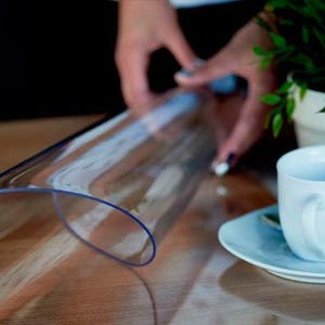 May include: A flexible, transparent, rectangular plastic sheet with rounded corners, positioned on a wooden surface next to a white ceramic coffee cup and saucer, with a small green plant in a white pot visible in the background.
