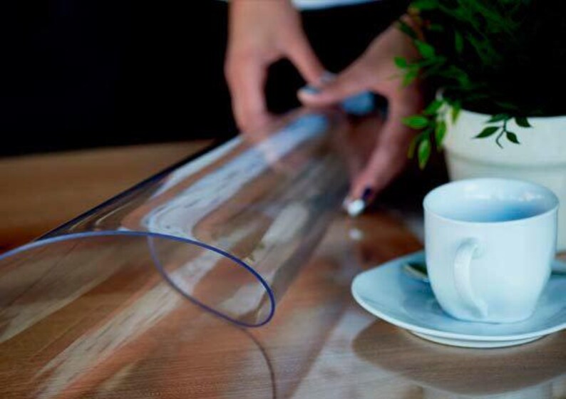 May include: A flexible, transparent, rectangular plastic sheet with rounded corners, positioned on a wooden surface next to a white ceramic coffee cup and saucer, with a small green plant in a white pot visible in the background.