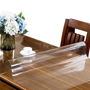 May include: A clear, flexible plastic table protector sheet laid over a dark brown wooden table. The transparent cover is smooth and glossy, partially unrolled to reveal the wood grain underneath, while a white cup and a vase of flowers sit nearby.
