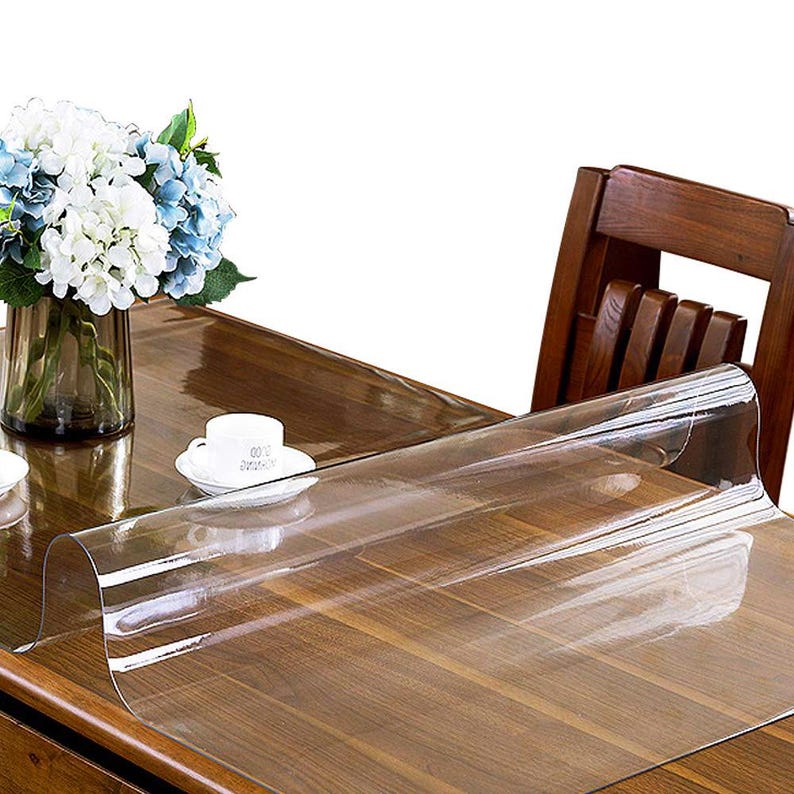 May include: A clear, flexible plastic table protector sheet laid over a dark brown wooden table. The transparent cover is smooth and glossy, partially unrolled to reveal the wood grain underneath, while a white cup and a vase of flowers sit nearby.