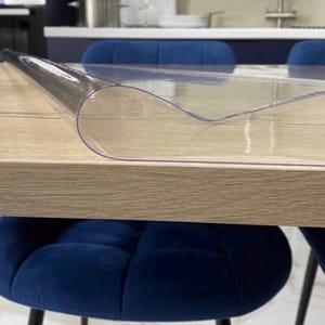 Może przedstawiać: Przezroczysta, elastyczna plastikowa mata ochronna na st&oacute;ł, leżąca płasko na drewnianym stole jadalnianym. Materiał jest przezroczysty, gładki i ma lekkie zawinięcie na krawędzi, co ukazuje jego cienką, giętką i wytrzymałą konstrukcję.