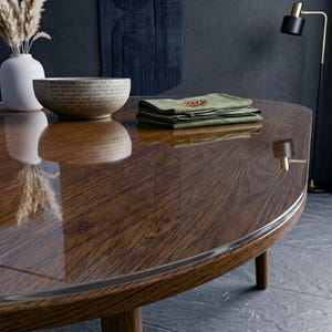 May include: A polished oval wooden coffee table with a dark brown grain pattern and a glass top. On the table sits a white ceramic bowl with printed text, a stack of folded olive green napkins with a small embroidered detail, and a white vase.