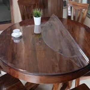 May include: A clear, circular plastic table protector mat laid over a round, dark brown wooden dining table. The transparent cover is partially lifted on one side, revealing the polished wood grain surface underneath. A small white cup and a potted plant sit on top.
