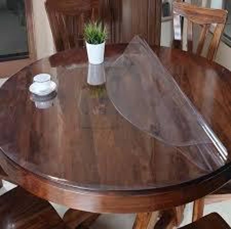 May include: A clear, circular plastic table protector mat laid over a round, dark brown wooden dining table. The transparent cover is partially lifted on one side, revealing the polished wood grain surface underneath. A small white cup and a potted plant sit on top.