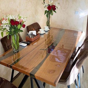 May include: A rectangular dining table featuring two natural wood slabs joined by a dark, translucent resin river inlay. The table has a glossy finish, black metal legs, and is surrounded by brown upholstered chairs with a bouquet of flowers.