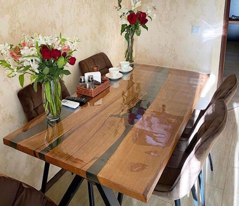 May include: A rectangular dining table featuring two natural wood slabs joined by a dark, translucent resin river inlay. The table has a glossy finish, black metal legs, and is surrounded by brown upholstered chairs with a bouquet of flowers.