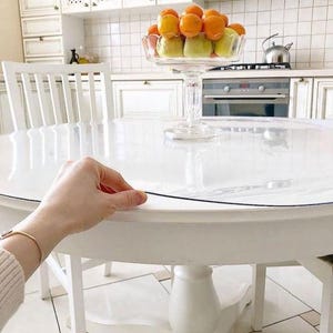 May include: A clear, circular plastic table protector cover resting on a white round wooden table. The transparent protective sheet has a smooth surface and is positioned to cover the tabletop, with a hand visible lifting the edge of the cover.