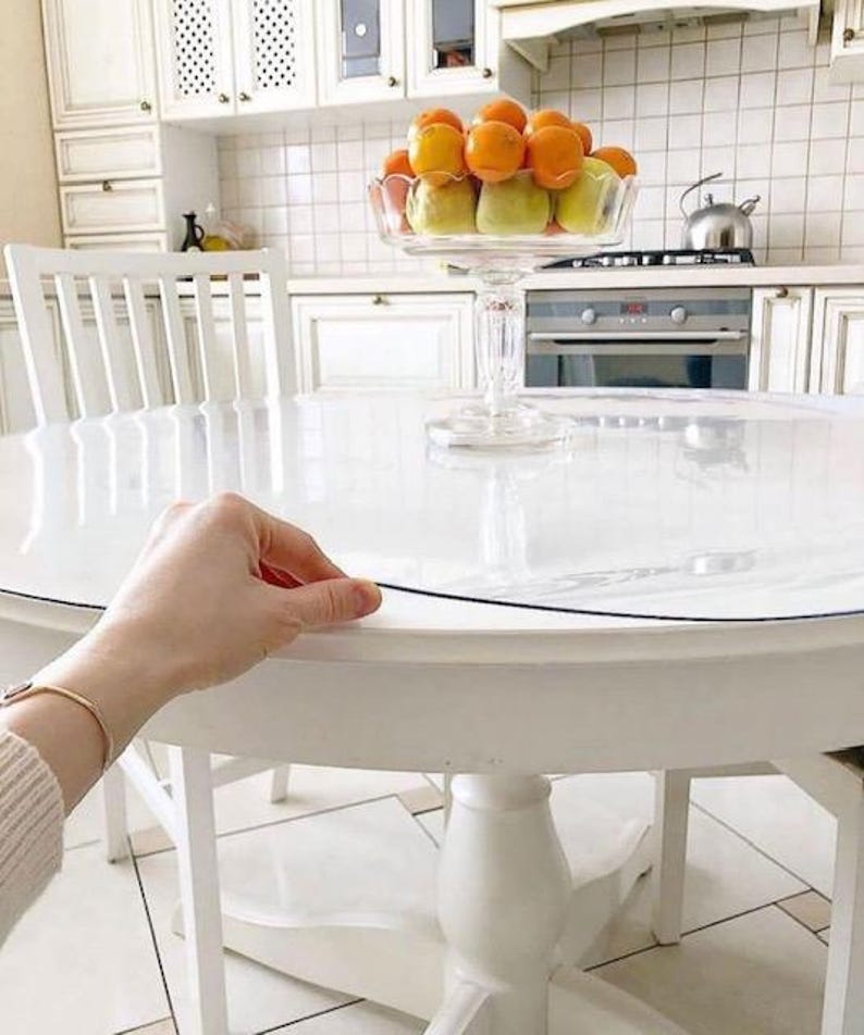 May include: A clear, circular plastic table protector cover resting on a white round wooden table. The transparent protective sheet has a smooth surface and is positioned to cover the tabletop, with a hand visible lifting the edge of the cover.