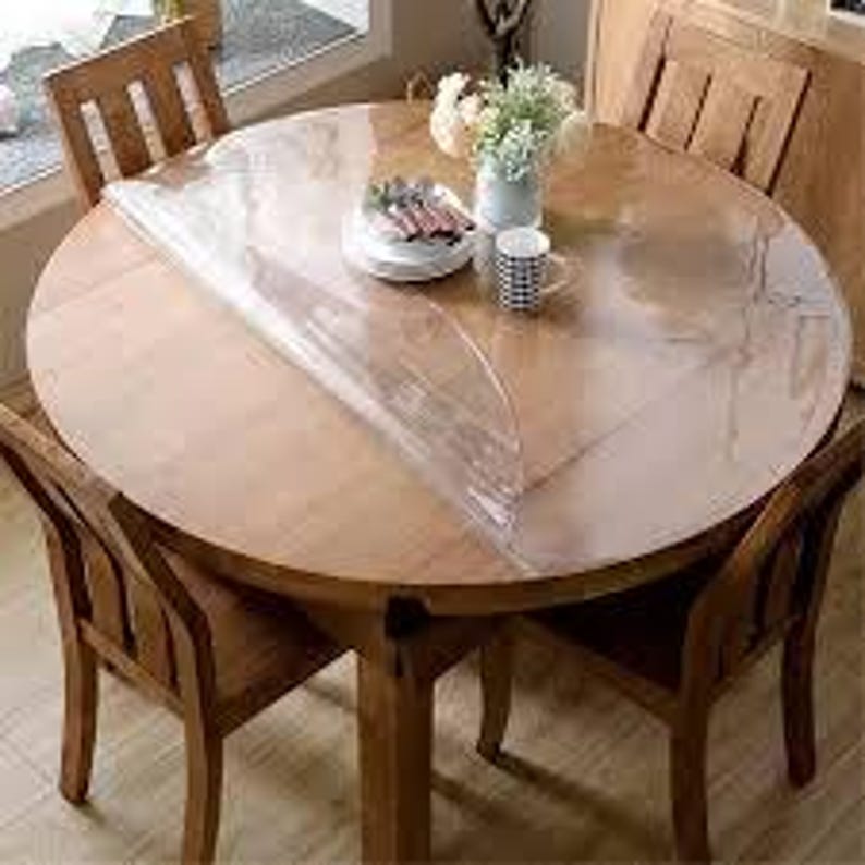 May include: A round, transparent plastic table protector sheet partially covering a circular wooden dining table. The clear, flexible cover lies flat over the wood surface, which is surrounded by wooden chairs and holds a vase and dishware.