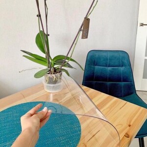 May include: A clear, flexible plastic table protector sheet being lifted by a hand, revealing a light wood grain table surface underneath with a round teal textured placemat and a potted orchid plant in a clear plastic pot.