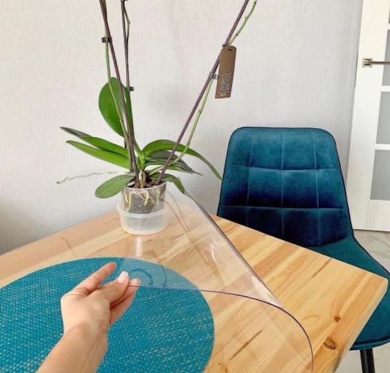 May include: A clear, flexible plastic table protector sheet being lifted by a hand, revealing a light wood grain table surface underneath with a round teal textured placemat and a potted orchid plant in a clear plastic pot.