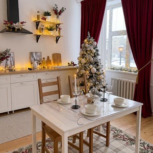 Könnte beinhalten: Ein Esstisch mit einer transparenten Schutzfolie, gedeckt mit weißen Schüsseln und Weingläsern. Der Raum ist mit einem geschmückten Weihnachtsbaum, dunkelroten Samtvorhängen, Holzstühlen und einer Küche mit weißen Schränken ausgestattet.