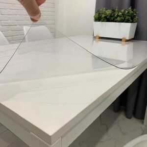 May include: A clear, flexible plastic table protector sheet being lifted from a white rectangular table. The transparent material is thin and smooth, designed to cover and protect the surface of the furniture while remaining visible.