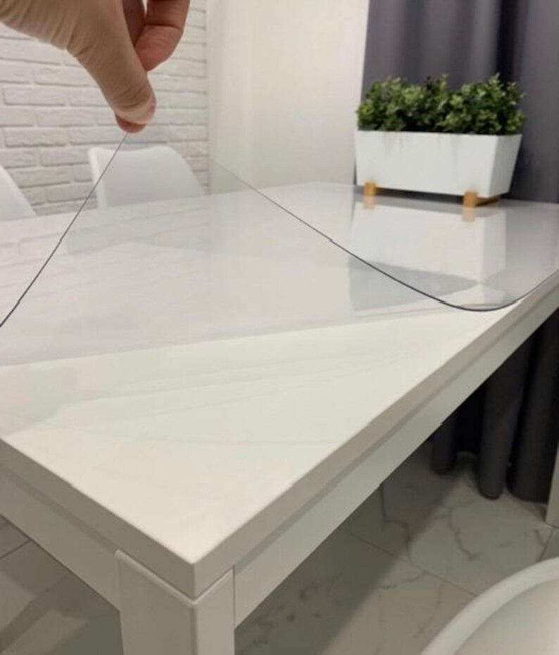 May include: A clear, flexible plastic table protector sheet being lifted from a white rectangular table. The transparent material is thin and smooth, designed to cover and protect the surface of the furniture while remaining visible.