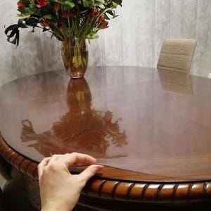 May include: A clear, flexible plastic table protector sheet laid over a dark brown wooden oval dining table. The protector is transparent, showing the wood grain underneath, and is positioned to cover the surface of the table completely.