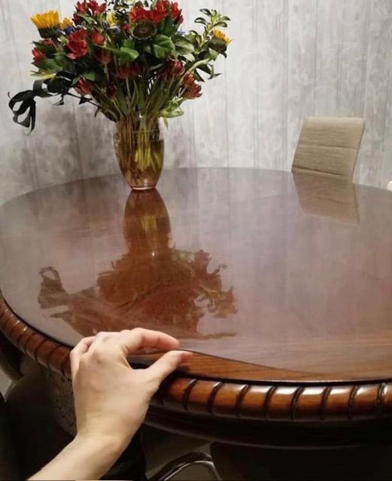 May include: A clear, flexible plastic table protector sheet laid over a dark brown wooden oval dining table. The protector is transparent, showing the wood grain underneath, and is positioned to cover the surface of the table completely.