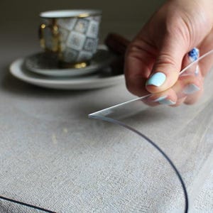 May include: A flexible, transparent plastic sheet protector resting on a textured light-colored surface. The clear material is lifted at one corner, revealing its smooth, pliable texture and thin profile while covering the underlying fabric surface.