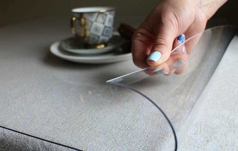 May include: A flexible, transparent plastic sheet protector resting on a textured light-colored surface. The clear material is lifted at one corner, revealing its smooth, pliable texture and thin profile while covering the underlying fabric surface.