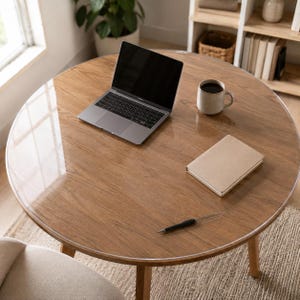 May include: A round wooden table with a clear protective cover, featuring a silver laptop, a beige ceramic mug filled with dark liquid, a closed beige notebook, and a black ballpoint pen placed on the smooth, reflective surface of the tabletop.