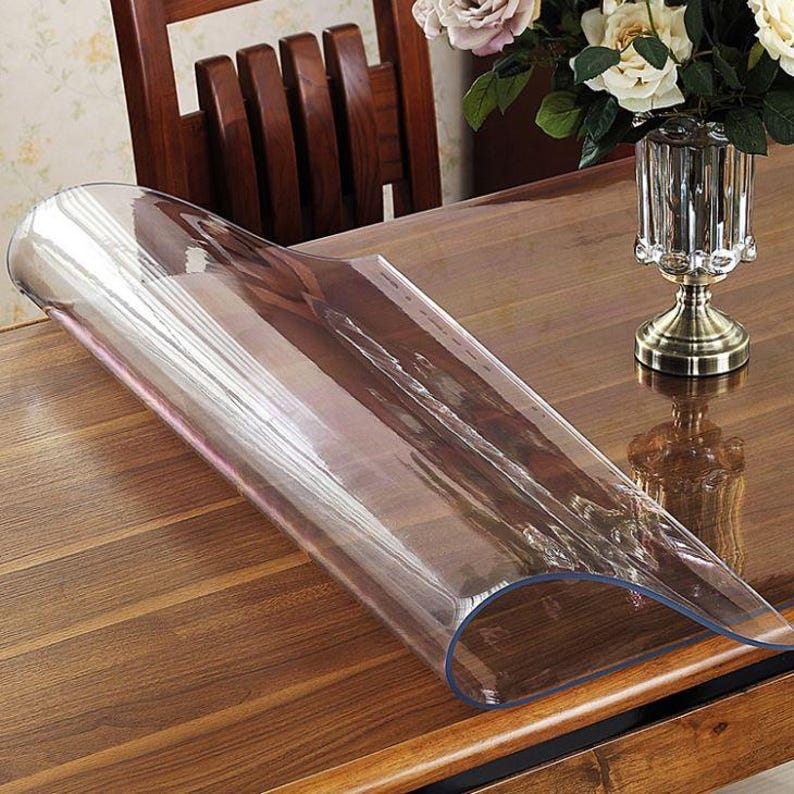 May include: A clear, flexible plastic table protector sheet partially rolled up on a polished wooden table surface. The transparent material reveals the wood grain underneath, and a glass vase with white flowers sits nearby on the table.