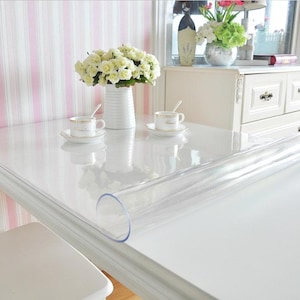 Op de afbeelding: Een doorzichtige plastic tafelkleed opgerold op een witte tafel. Op de tafel staan twee kopjes en schotels, een witte vaas met bloemen en een roze en witte gestreepte muur op de achtergrond.