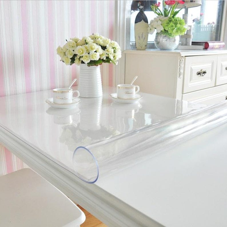 May include: A clear, flexible plastic table protector sheet laid flat over a white table surface. The transparent material is partially rolled up at one end, revealing its smooth texture. Two white ceramic cups and a vase of white flowers sit on top.