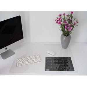 May include: A clear rectangular desk mat placed on a white surface, featuring a 2022 calendar print in the bottom right corner. The mat is positioned under a white computer keyboard, a white computer mouse, a computer monitor, and a grey vase.