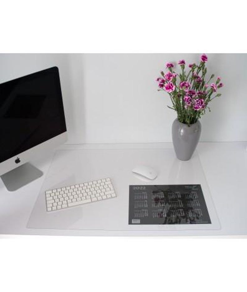 May include: A clear rectangular desk mat placed on a white surface, featuring a 2022 calendar print in the bottom right corner. The mat is positioned under a white computer keyboard, a white computer mouse, a computer monitor, and a grey vase.