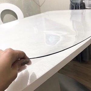 May include: A clear, flexible plastic protective cover sheet resting on top of a white rectangular table. The thin, transparent material is lifted slightly at the edge to demonstrate its pliable texture and smooth, glossy surface finish.
