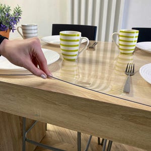 May include: A clear, flexible plastic table protector sheet laid flat over a light-toned wooden dining table. The transparent cover features a smooth surface, allowing the wood grain to remain visible while protecting the tabletop from spills.
