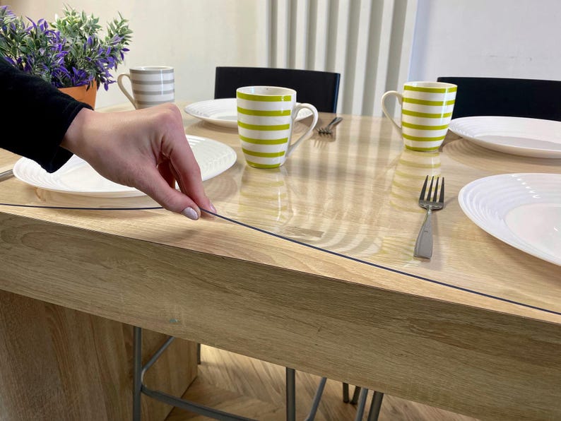 May include: A clear, flexible plastic table protector sheet laid flat over a light-toned wooden dining table. The transparent cover features a smooth surface, allowing the wood grain to remain visible while protecting the tabletop from spills.