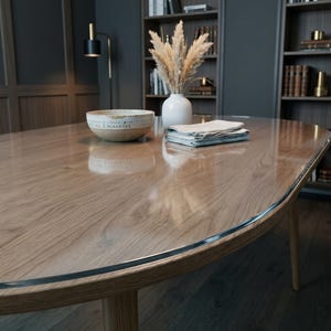 Könnte beinhalten: Ein ovaler Holzesstisch mit einer klaren Glasplatte. Auf dem Tisch stehen eine beige Keramikschale mit Text, eine weiße Vase mit getrocknetem Pampasgras und ein Stapel gefalteter, heller Stoffservietten. Der Tisch hat eine glatte Oberfläche.