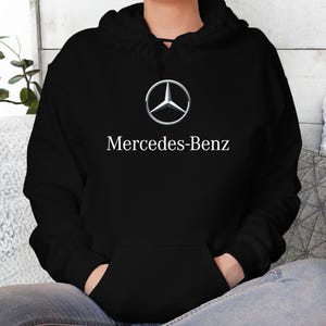 May include: Black hooded sweatshirt with a front pocket and the Mercedes-Benz logo in silver and white. The brand name is printed below the logo in white. The sweatshirt is made of a soft material.