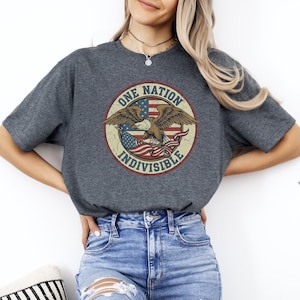 May include: Dark gray t-shirt featuring a distressed circular graphic with an eagle, American flag, and the words "ONE NATION INDIVISIBLE". The shirt is made of a soft material and has short sleeves.