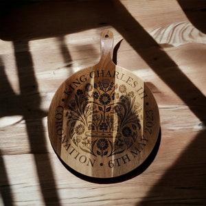 May include: A round wooden serving board with a handle. The board is decorated with a floral design and the text "King Charles III Coronation 6th May 2023".