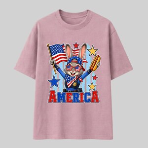 Puede incluir: Una camiseta rosa claro con un conejo de dibujos animados vestido de polic&iacute;a, con gafas de sol estrelladas y un pa&ntilde;uelo. El conejo sostiene una bandera estadounidense y un perrito caliente. La palabra AMERICA est&aacute; impresa en letras rojas y azules.