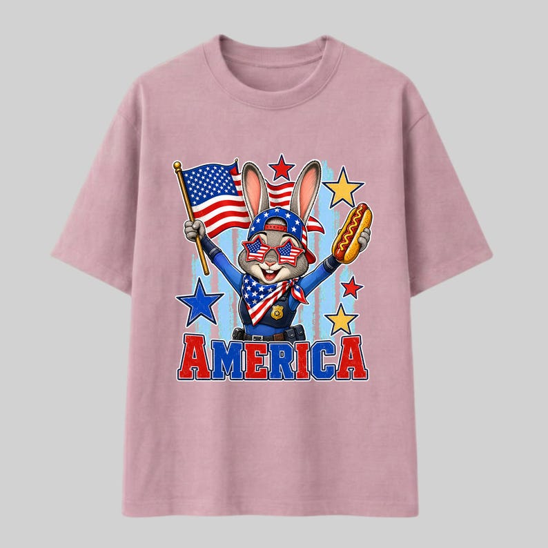 Puede incluir: Una camiseta rosa claro con un conejo de dibujos animados vestido de polic&iacute;a, con gafas de sol estrelladas y un pa&ntilde;uelo. El conejo sostiene una bandera estadounidense y un perrito caliente. La palabra AMERICA est&aacute; impresa en letras rojas y azules.
