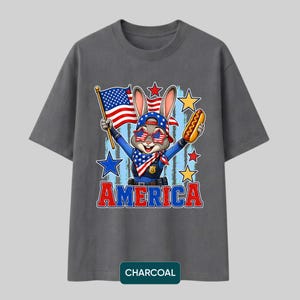 Puede incluir: Una camiseta gris carb&oacute;n con un conejo de dibujos animados con uniforme de polic&iacute;a, un pa&ntilde;uelo con patr&oacute;n de bandera y gafas de sol en forma de estrella. El conejo sostiene una bandera estadounidense y un perrito caliente. La palabra AMERICA est&aacute; en rojo y azul.